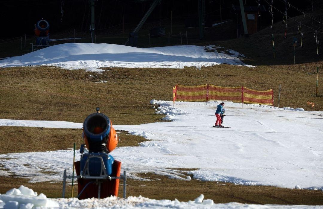 Eine Schneekanone steht vor einer Kunstschnee-Piste auf der eine Frau mit Kind fahren, daneben braun-grüne Wiesen.