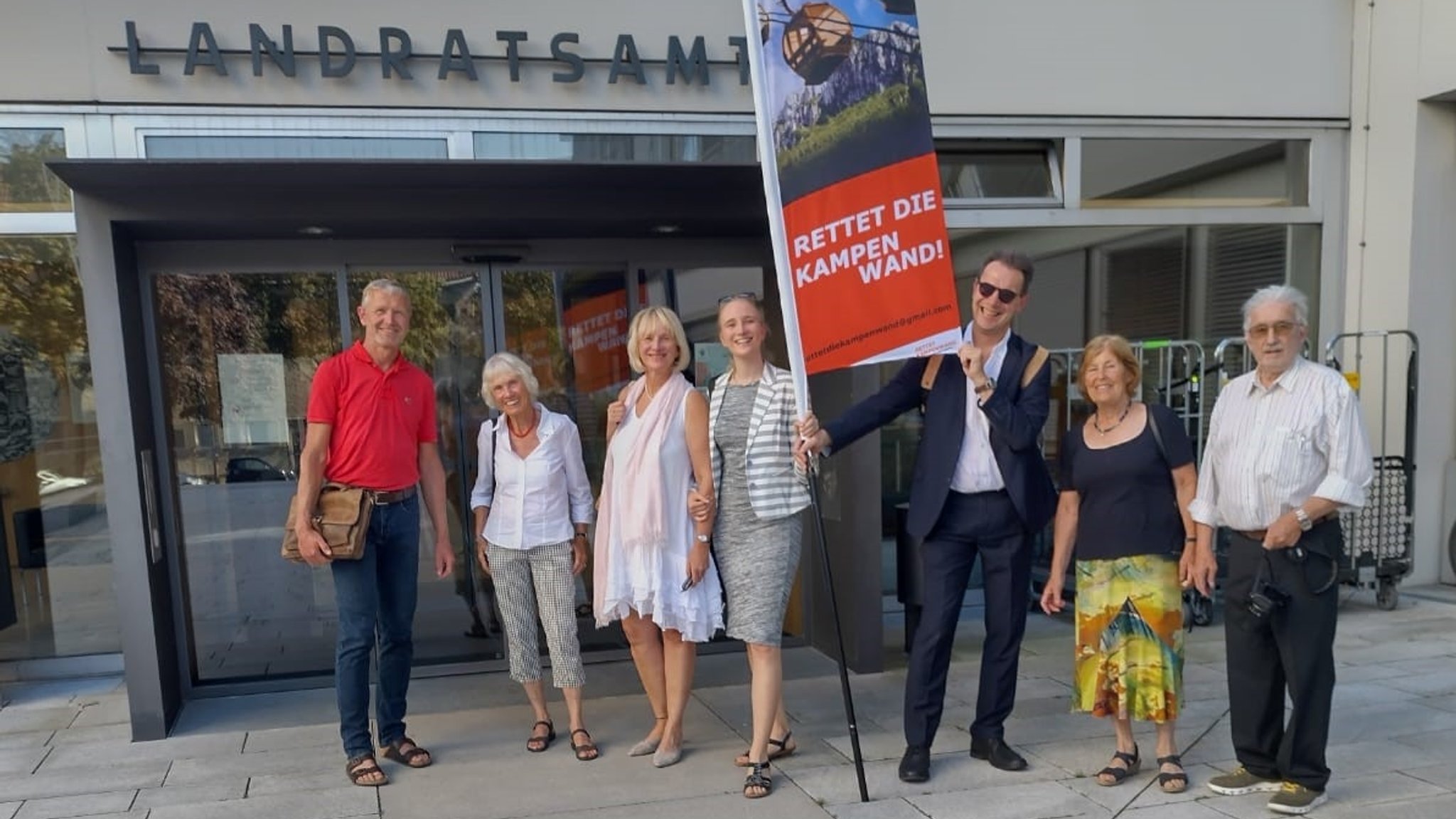 Vertreter von Bürgerinitiative,  vom Bund Naturschutz und vom Verein zum Schutz der Bergwelt stehen mit einer Fahne mit der Aufschrift "Rettet die Kampenwand!" vor dem Rosenheimer Landratsamt