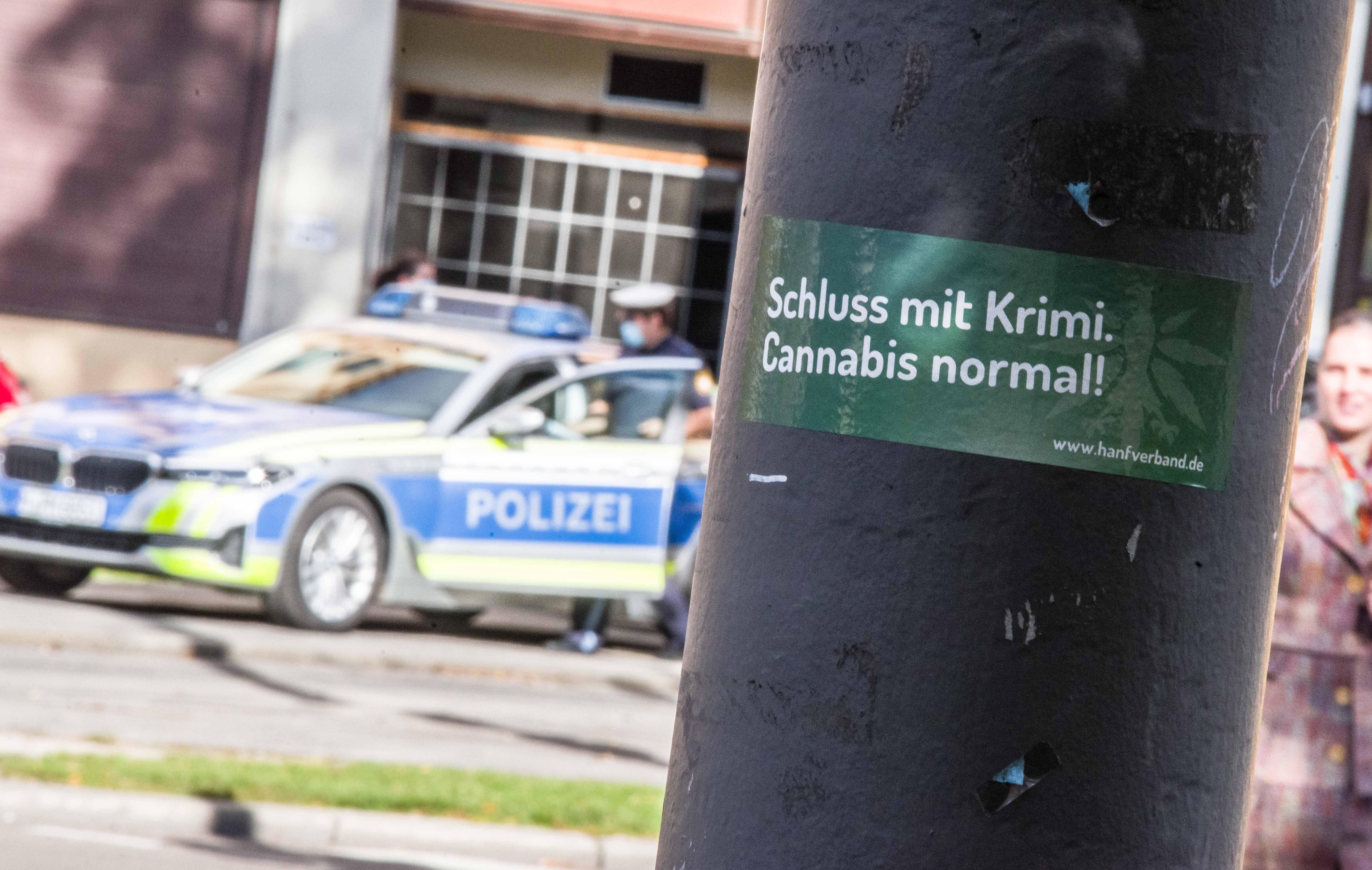 Ein Cannabis-Aufkleber an einer Säule in München, dahinter steht die Polizei 