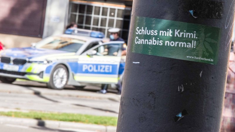 Ein Cannabis-Aufkleber an einer Säule in München, dahinter steht die Polizei 