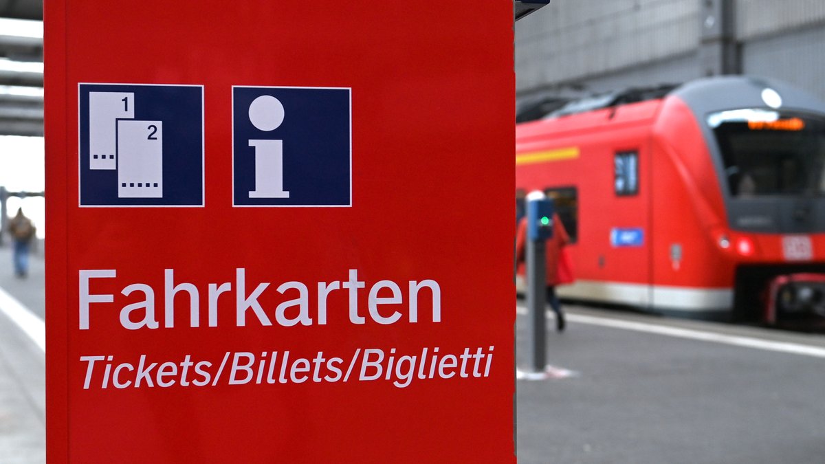 Ein Fahrkartenautomat der Deutschen Bahn vor einem Bahngleis, an dem ein Regionalzug wartet.