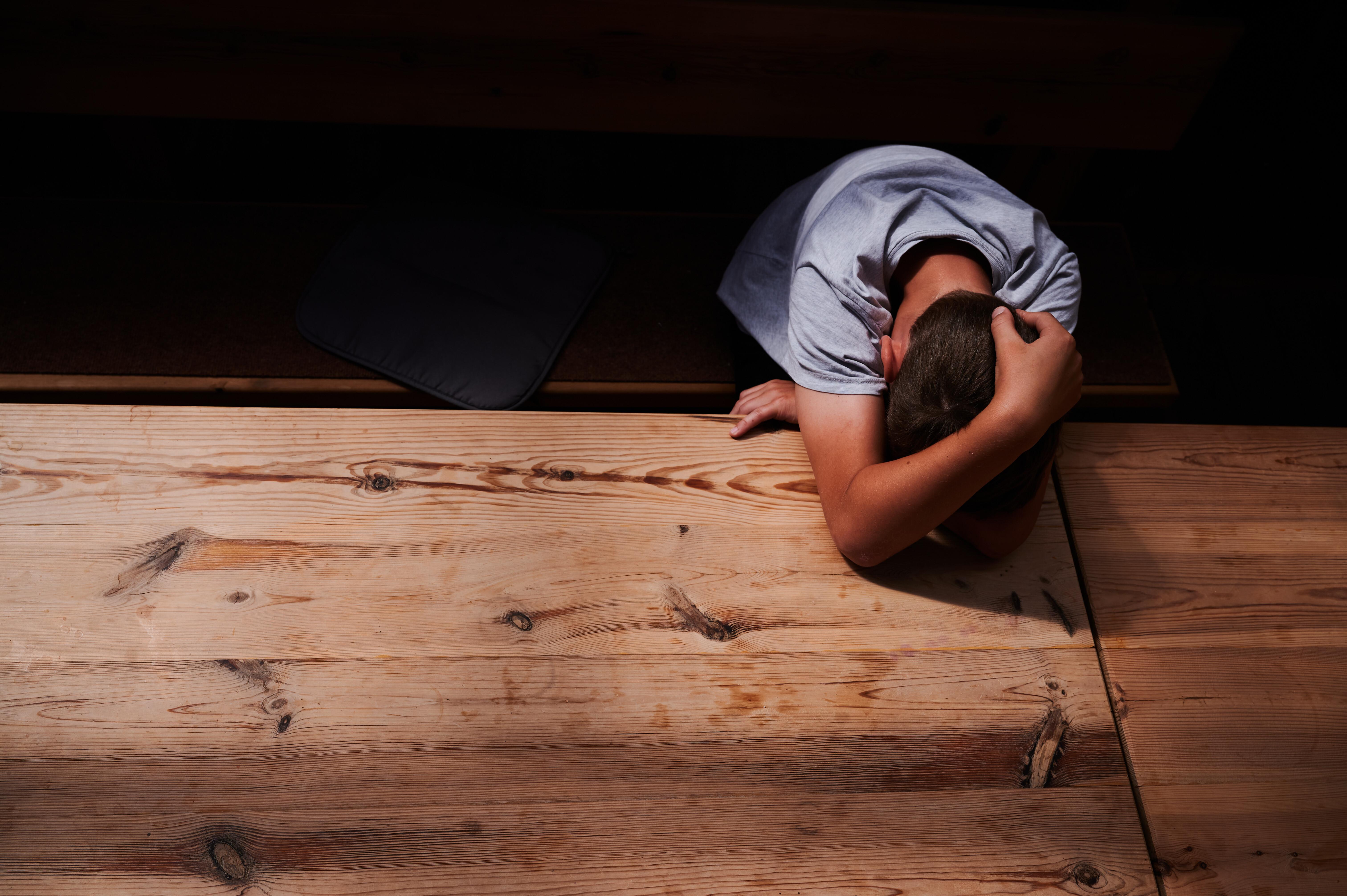 Ein Jugendlicher versteckt sein Gesicht unter seinen Armen und liegt mit dem Kopf auf einem Tisch (gestellte Szene) (Archiv- und Symbolbild)