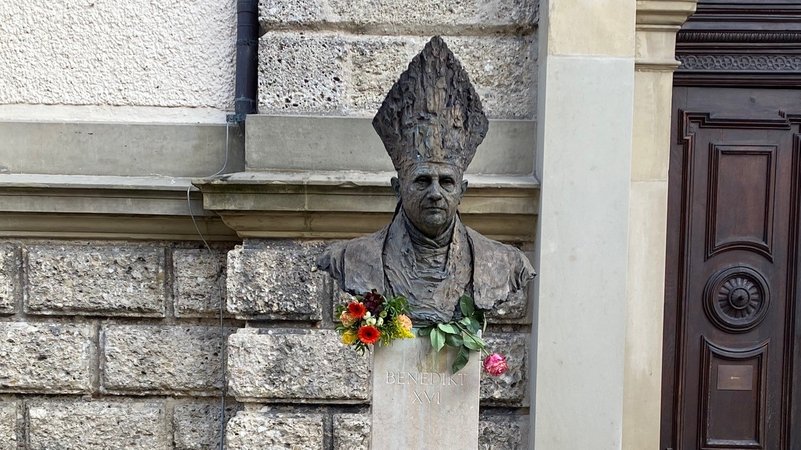 Die Büste von Benedikt XVI. vor der Kirchenmauer der katholischen Stadtpfarrkirche St. Oswald in Traunstein. Darunter liegen frische Blumen.
