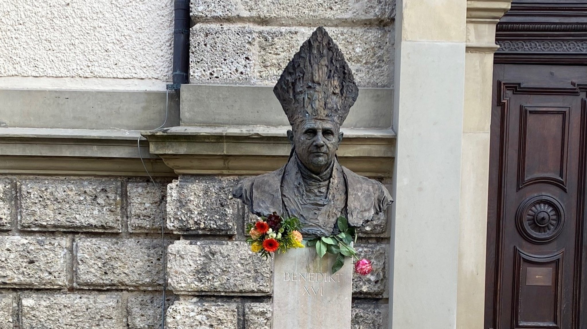 Die Büste von Benedikt XVI. vor der Kirchenmauer der katholischen Stadtpfarrkirche St. Oswald in Traunstein. Darunter liegen frische Blumen.