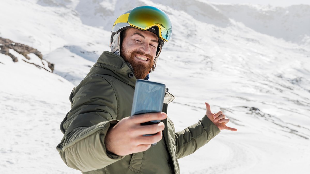 Lachender Mann im Ski-Anzug macht mit Smartphone ein Selfie vor Bergkulisse. 