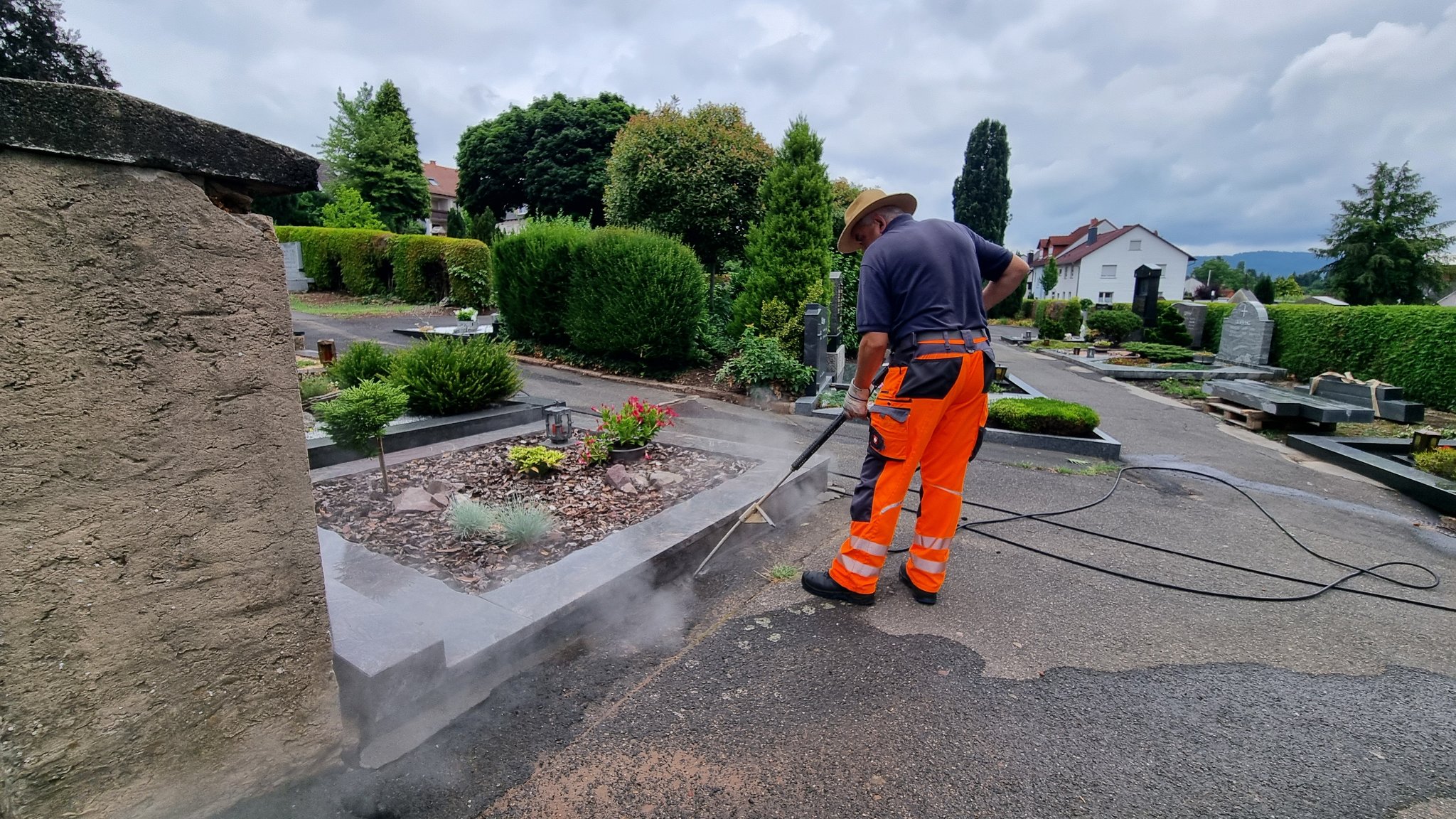 Auf dem Foto ist ein Mitarbeiter des Bauhofs auf einem Friedhof zu sehen. Er trägt orangefarbene Warnkleidung und reinigt mit einem Hochdruckreiniger den Asphalt entlang einer Grabumrandung. Wasserdampf steigt dabei vom Boden auf. Im Hintergrund sind gepflegte Gräber, Hecken, Bäume und Wohnhäuser unter einem bewölkten Himmel zu erkennen. Die Aufnahme verdeutlicht die Arbeiten zur Reinigung oder Bekämpfung von Ameisenbefall auf dem Friedhof. | Bild: BR / Albrecht Rauh Auf dem Foto ist ein Mitarbeiter des Bauhofs auf einem Friedhof zu sehen. Er trägt orangefarbene Warnkleidung und reinigt mit einem Hochdruckreiniger den Asphalt entlang einer Grabumrandung. Wasserdampf steigt dabei vom Boden auf. Im Hintergrund sind gepflegte Gräber, Hecken, Bäume und Wohnhäuser unter einem bewölkten Himmel zu erkennen. Die Aufnahme verdeutlicht die Arbeiten zur Reinigung oder Bekämpfung von Ameisenbefall auf dem Friedhof.