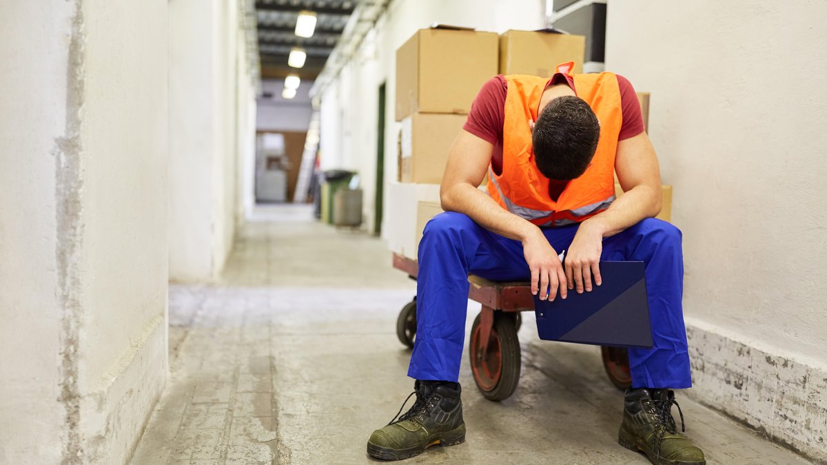 Arbeiter sitzt erschöpft auf einem Schubwagen im Warenlager wegen Überstunden.