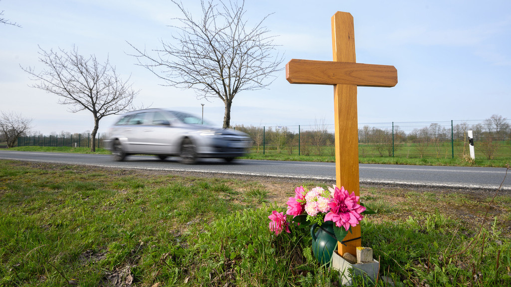 Ein Holzkreuz mit Blumen steht an einer Landstraße, ein Auto fährt im Hintergrund vorbei.