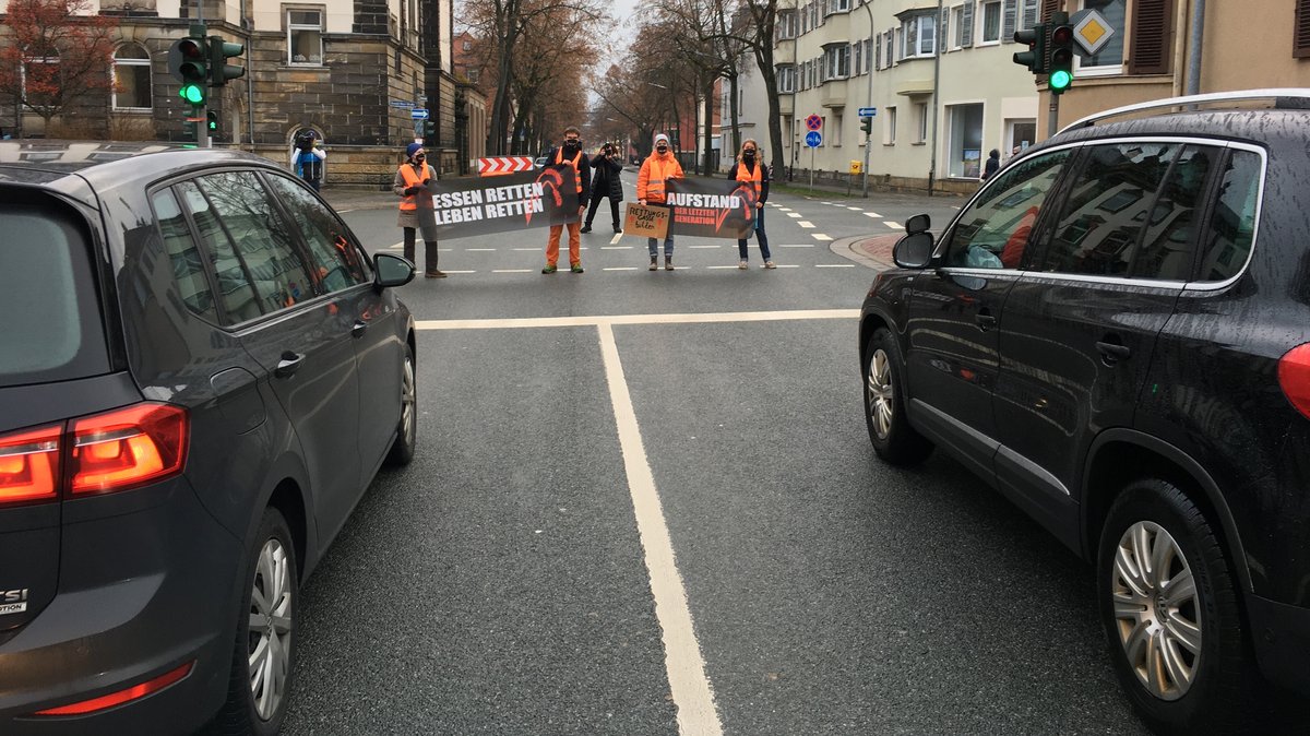 Vier Personen mit orangenen Warnwesten stehen auf einer Bayreuther Straßenkreuzung mit Plakaten und behindern den Verkehr.