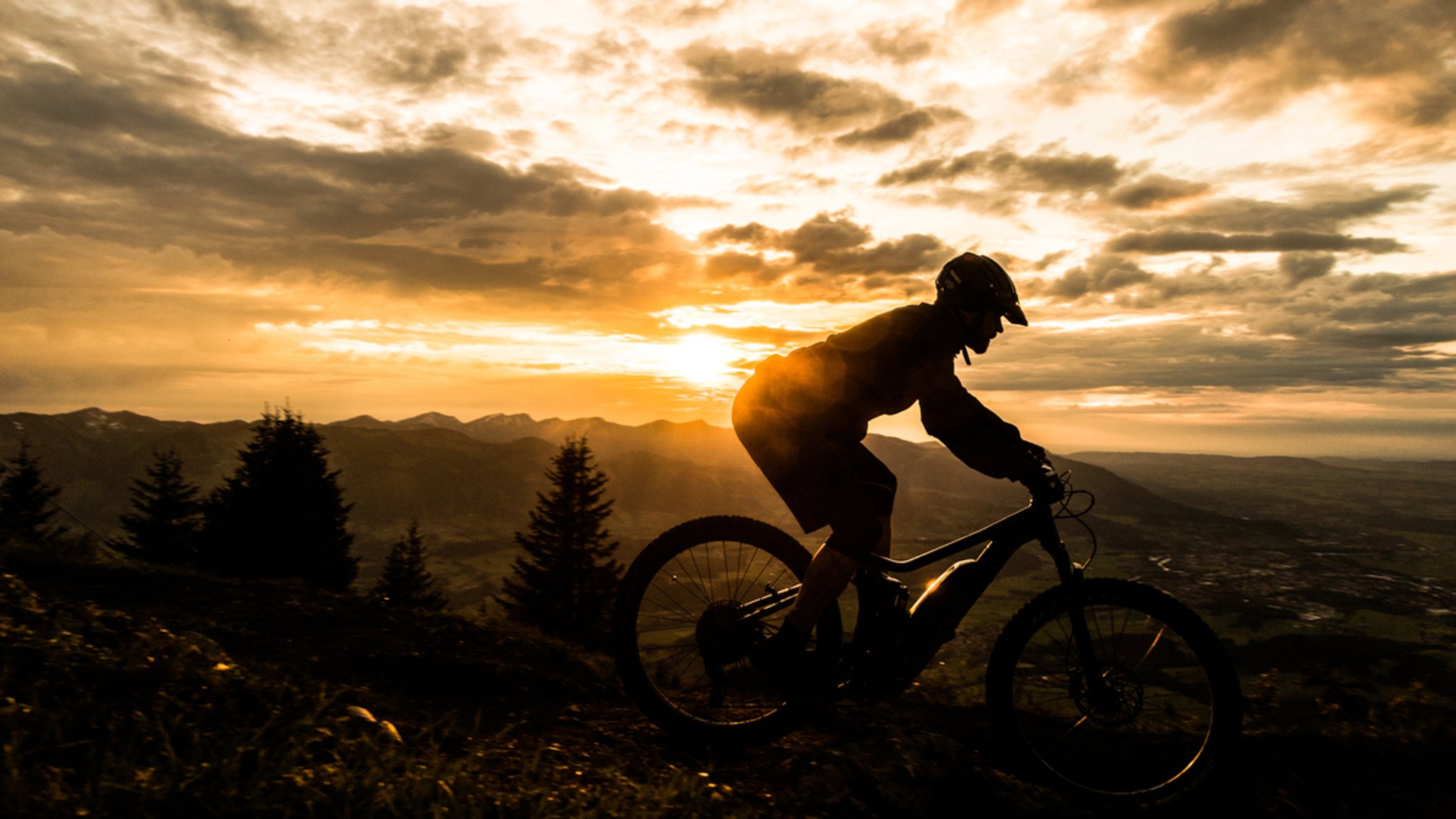 Ein Mann auf einem Mountainbike fährt in luftiger Höhe. Hinter ihm ist eine Aussicht mit Bergen zu sehen, dahinter geht die Sonne unter. 