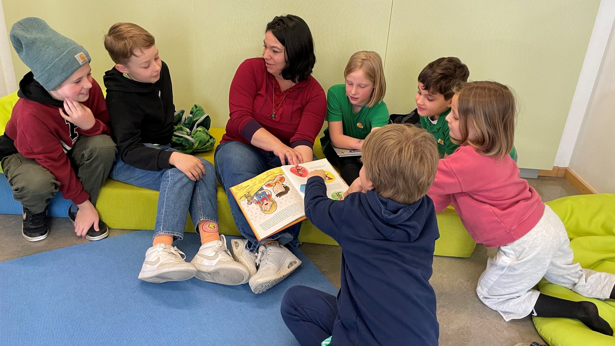 Eine Pädagogin mit einem Buch ist umringt von Grundschulkindern. 