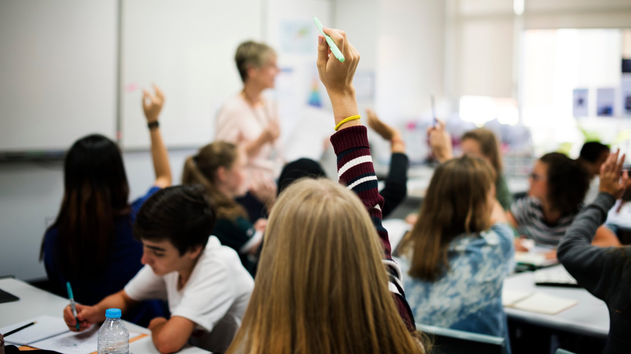 Schülerinnen und Schüler sitzen in einem Klassenzimmer. An der Tafel steht eine Lehrerin. 