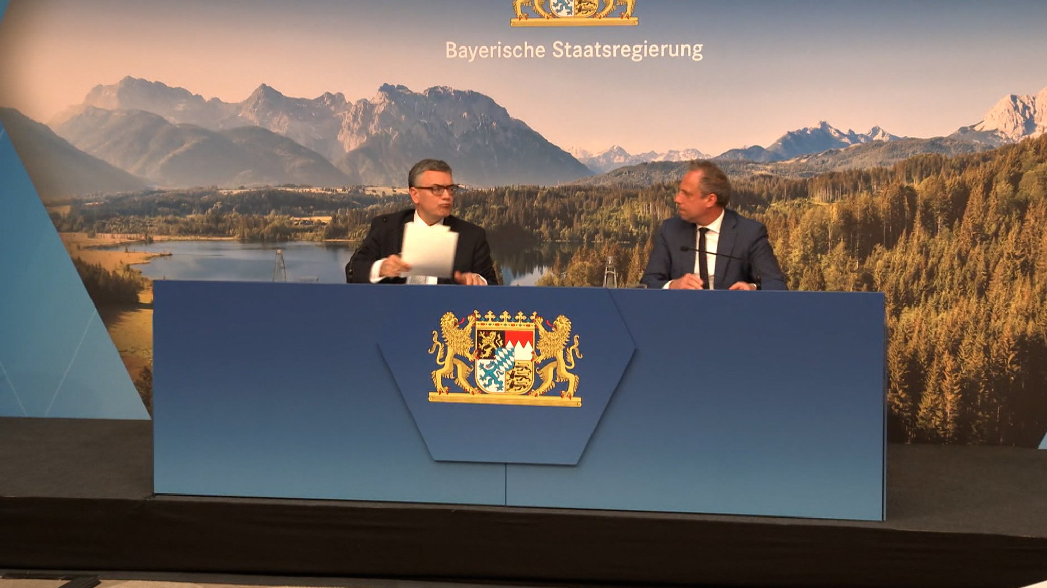 Zwei Männer sitzen hinter einem blauen Pult bei einer Pressekonferenz.