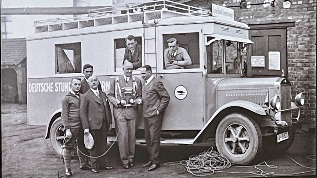 Bild eines Übertragungswagens des Bayerischen Rundfunks (Deutsche Stunde in Bayern), davor stehen mehrere Männer.