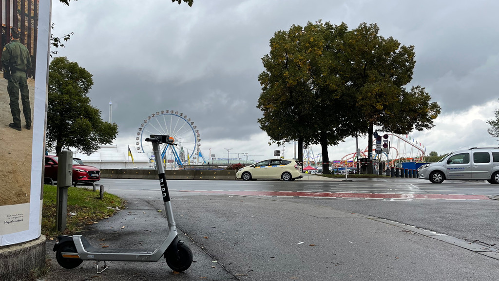 Ein silberner E-Scooter steht am Straßenrand. Im Hintergrund fahren Ein Taxi und zwei Autos vorbei. Außerdem sind hinter der Straße ein Riesenrad und andere Fahrgeschäfte des Oktoberfestes zu sehen,