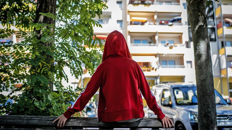 Ein Jugendlicher in roter Kapuzenjacke sitzt auf einem Zaun vor einem Hochhaus.