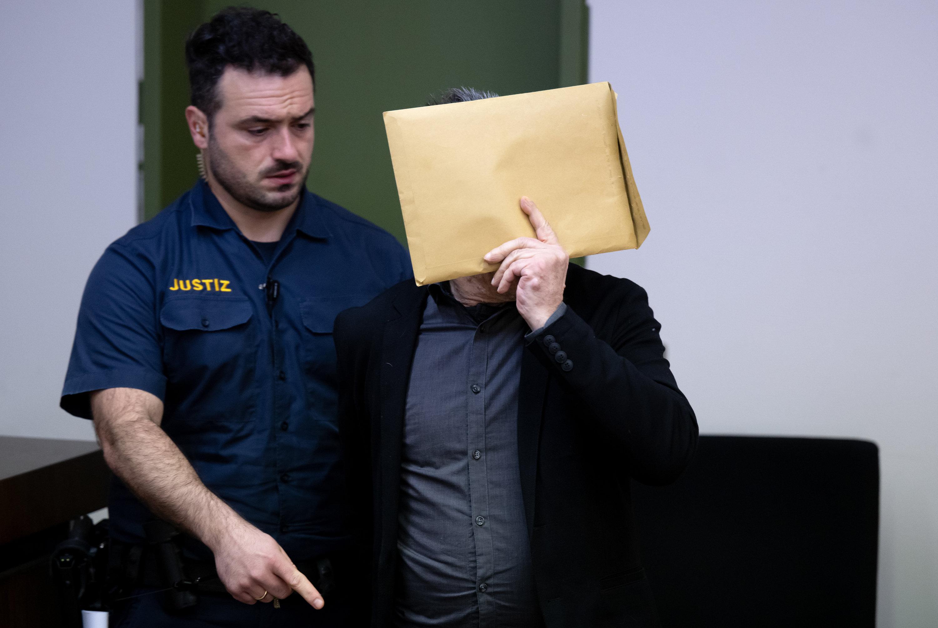 Der Angeklagte verdeckt sein Gesicht mit einem Briefumschlag; neben ihm steht ein Justizbeamter.