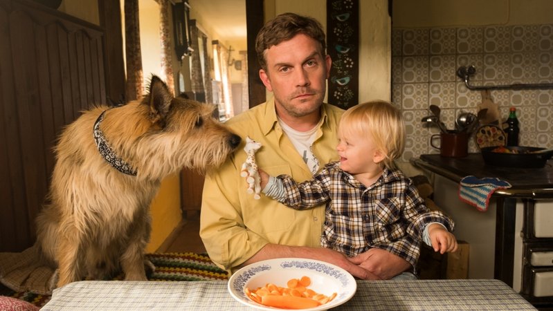 Filmszene aus "Leberkäsjunkie": Schauspieler Sebastian Bezzel als Eberhofer sitzt am Küchentisch, mit Kleinkind am Schoß und Hund