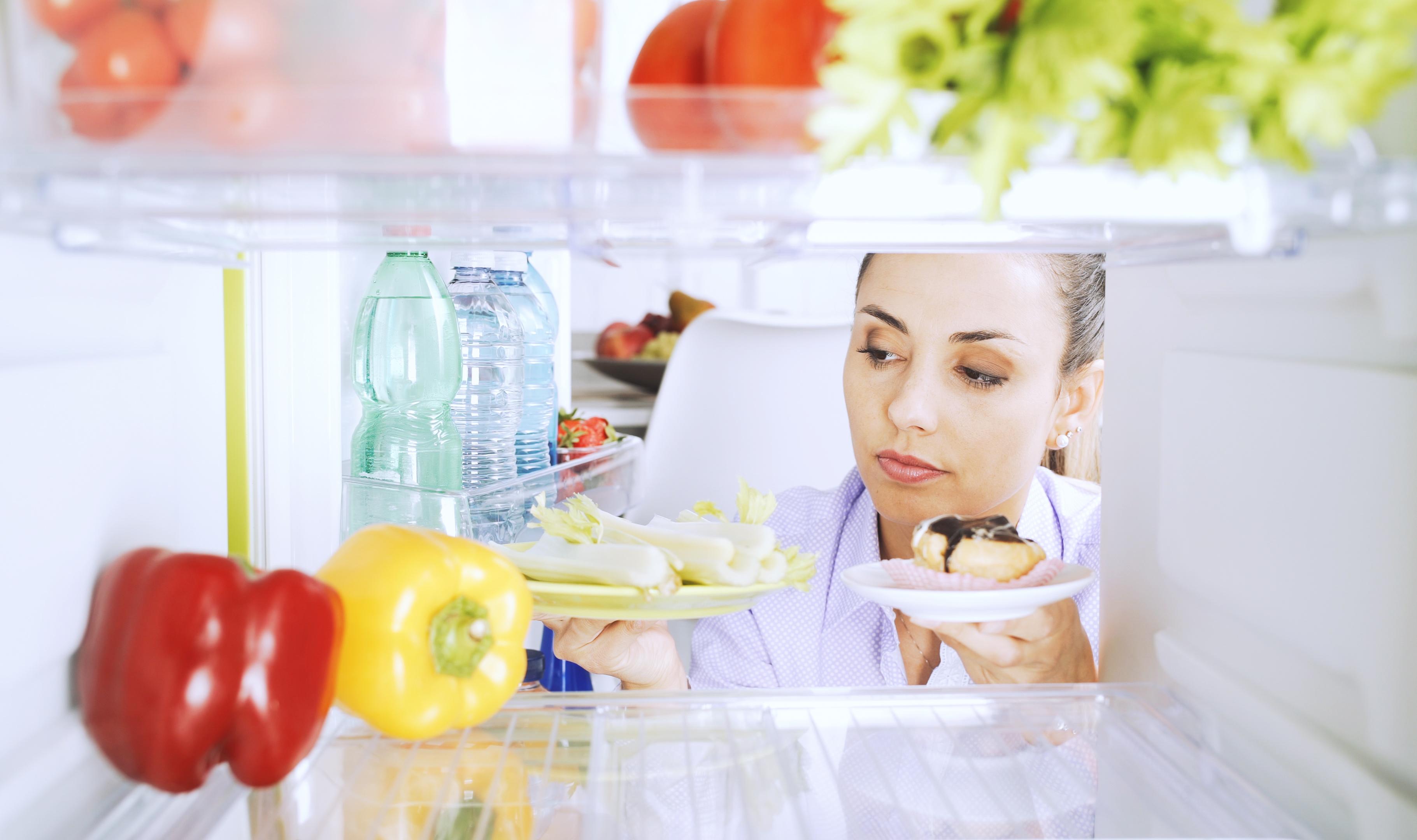 Eine junge Frau blickt sorgenvoll in einen Kühlschrank, der mit Obst, Gemüse und Süßigkeiten gefüllt ist