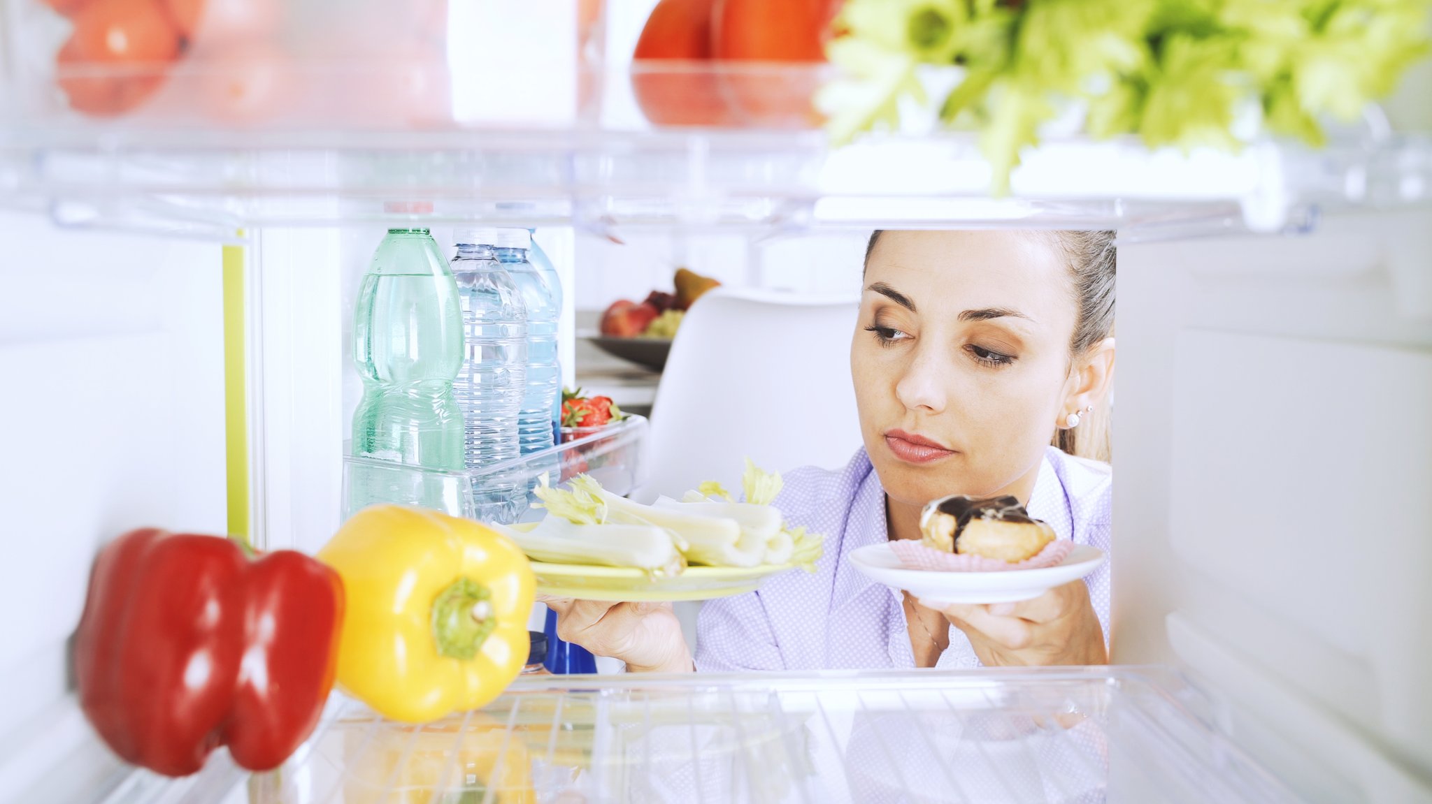 Eine junge Frau blickt sorgenvoll in einen Kühlschrank, der mit Obst, Gemüse und Süßigkeiten gefüllt ist