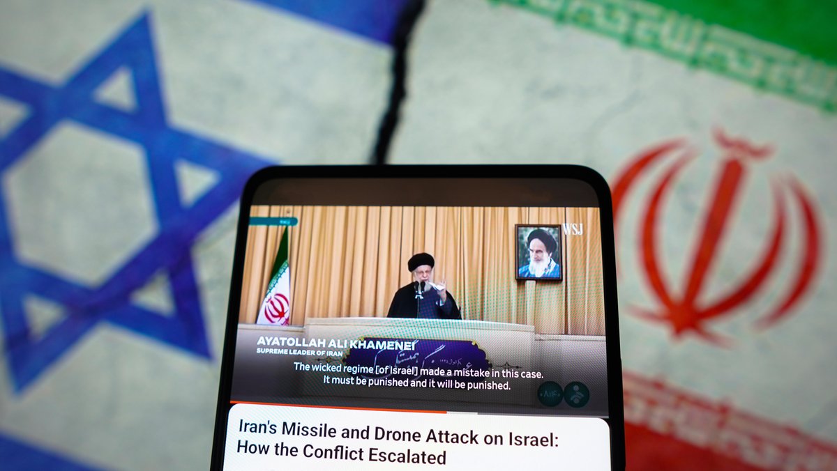 Collage: Israel und Iran-Flagge, davor ein Handy mit aktuellen News über eine Rede von Irans Revolutionsführer Ayatollah Ali Chamenei.