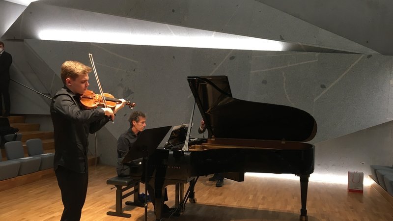 Zwei Männer konzertieren mit Geige und Piano in einem Konzertsaal, dessen Wände aus überdimensional großen Granitsplittern bestehen.