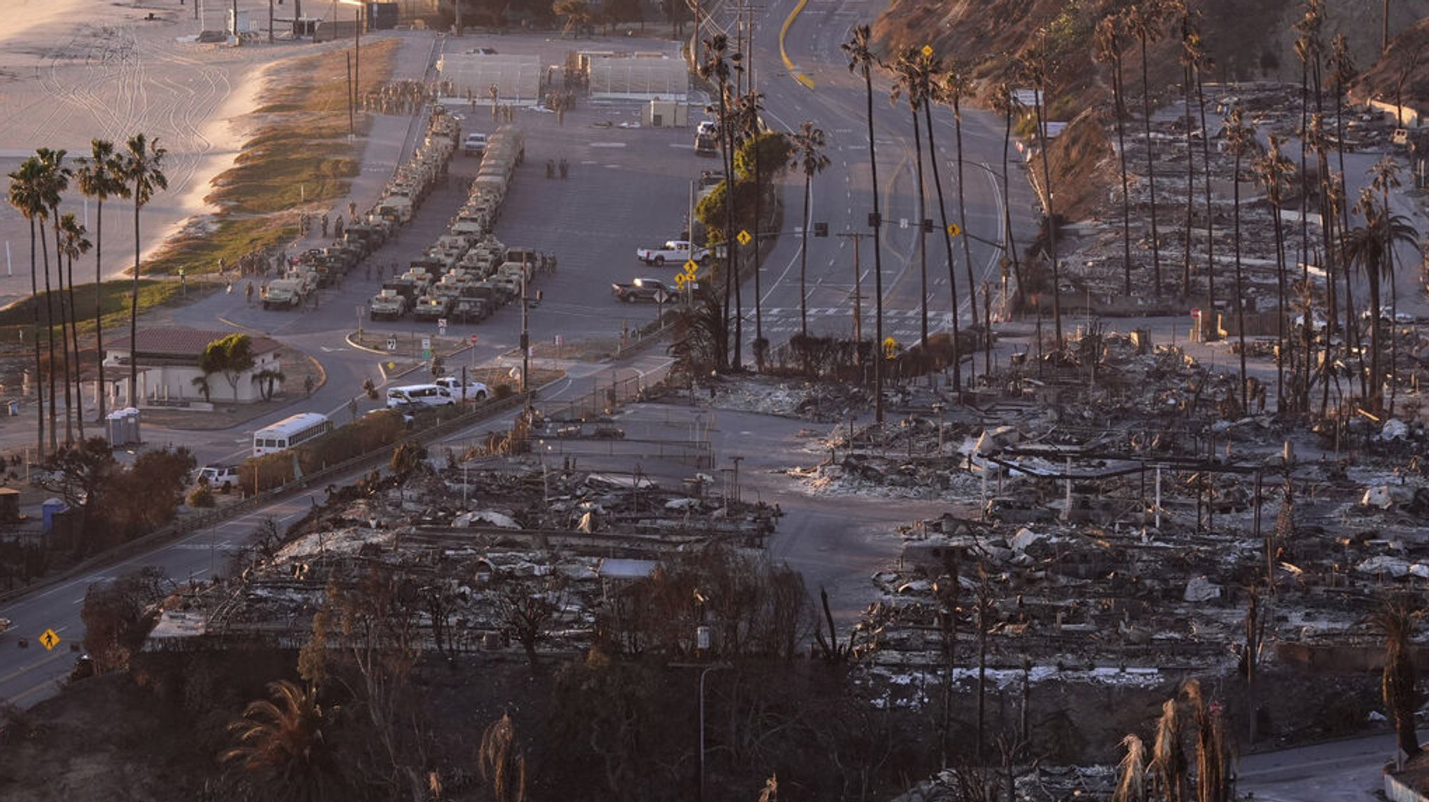 12.01.2025, USA, Los Angeles: Eine ausgebrannte Nachbarschaft ist entlang des Pacific Coach Highway nach dem Palisades-Feuer zu sehen, während sich mehrere Fahrzeuge der Nationalgarde am Strand aufstellen, im Stadtteil Pacific Palisades von Los Angeles. Foto: Mark J. Terrill/AP/dpa +++ dpa-Bildfunk +++