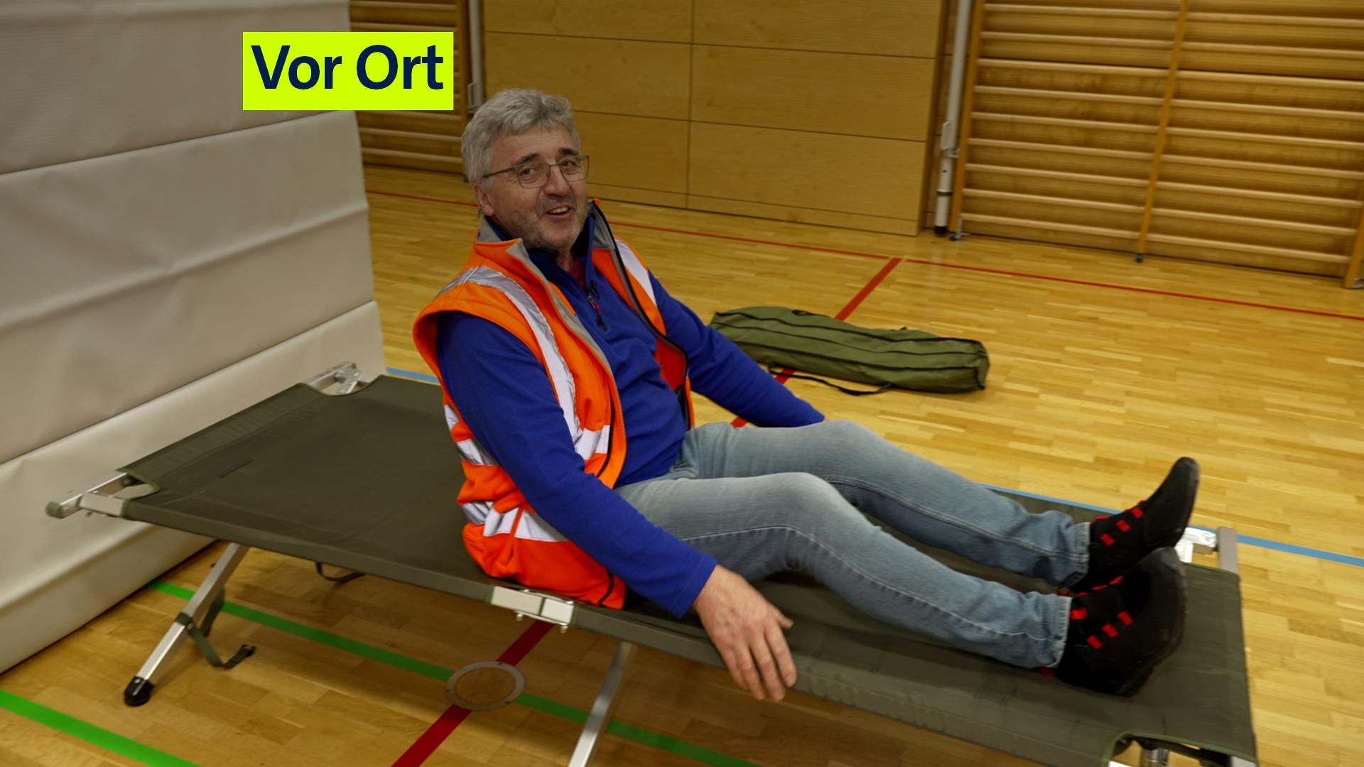 Ein Mann mit Warnweste, Pullover und Jeans auf einer einfachen Liege.