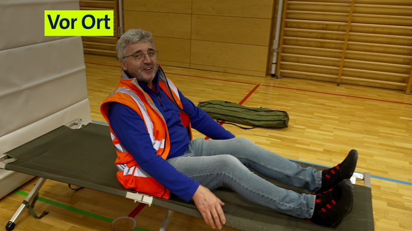 Ein Mann mit Warnweste, Pullover und Jeans auf einer einfachen Liege. | Bild: BR / Manon Hardenberg Ein Mann mit Warnweste, Pullover und Jeans auf einer einfachen Liege.