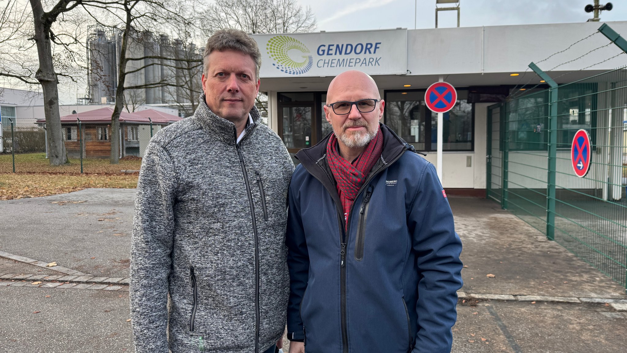 Zwei Männer stehen vor einem Zaun und schauen in die Kamera. Hinter ihnen sind chemische Anlagen und ein Schild mit der Aufschrift "Gendorf Chemiepark" zu sehen.