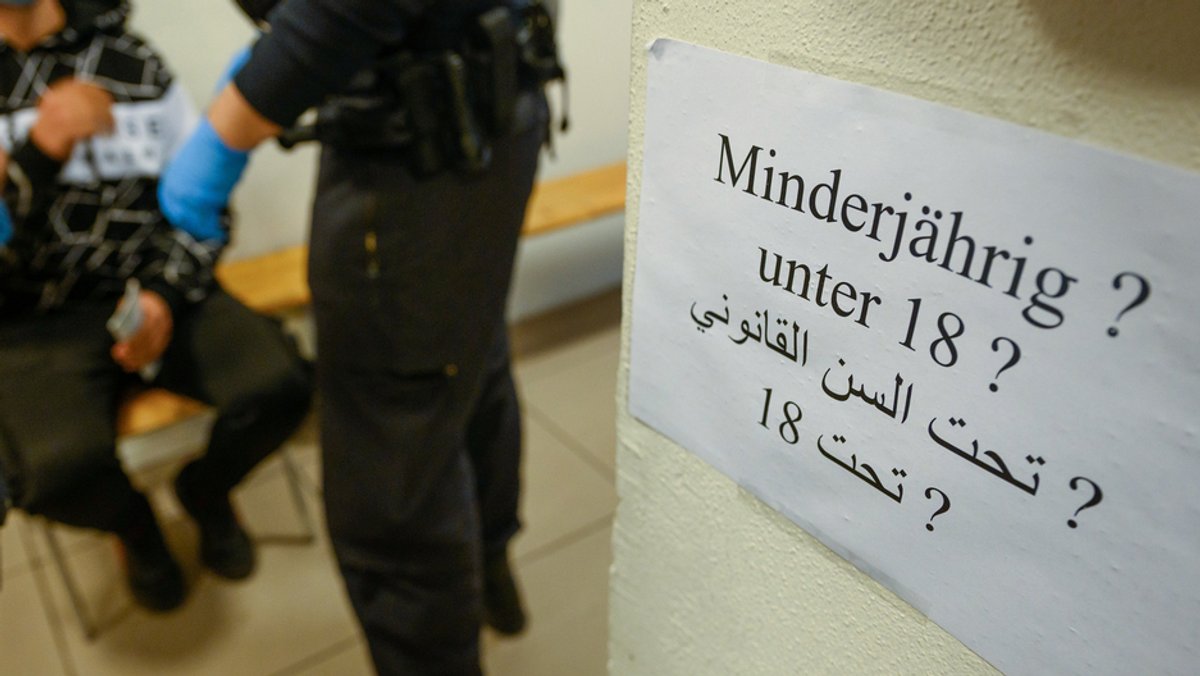 Auf einem Zettel steht auf deutsch und arabisch: "Minderjährig? Unter 18?". Im Hintergrund sieht man einen Polizisten und eine männliche Person auf einem Stuhl.