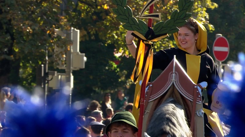 "Münchner Kindl" Franziska Inselkammer im schwarz-gelben Gewand winkt beim Trachten- und Schützenzug von ihrem Pferd. | Bild: Bayerischer Rundfunk 2024 "Münchner Kindl" Franziska Inselkammer im schwarz-gelben Gewand winkt beim Trachten- und Schützenzug von ihrem Pferd.