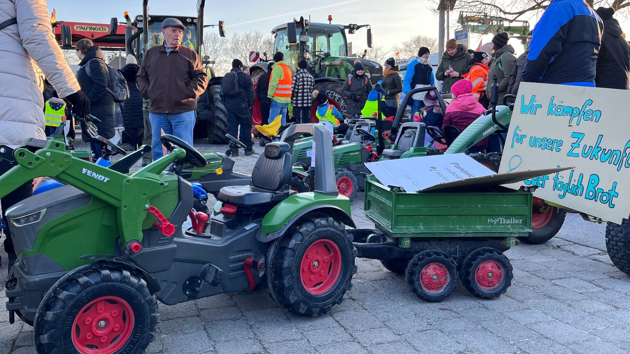 Ein grüner Tretschlepper für Kinder mit Anhänger steht in einer Menschenmenge. Protestplakate von Landwirten sind daran befestigt.