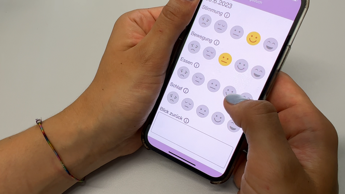 Jemand hält ein Handy in der Hand. Auf dem Display sind Emojis zum Thema Stimmung, Bewegung, Essen, Schlaf und Ernährung auszuwählen.