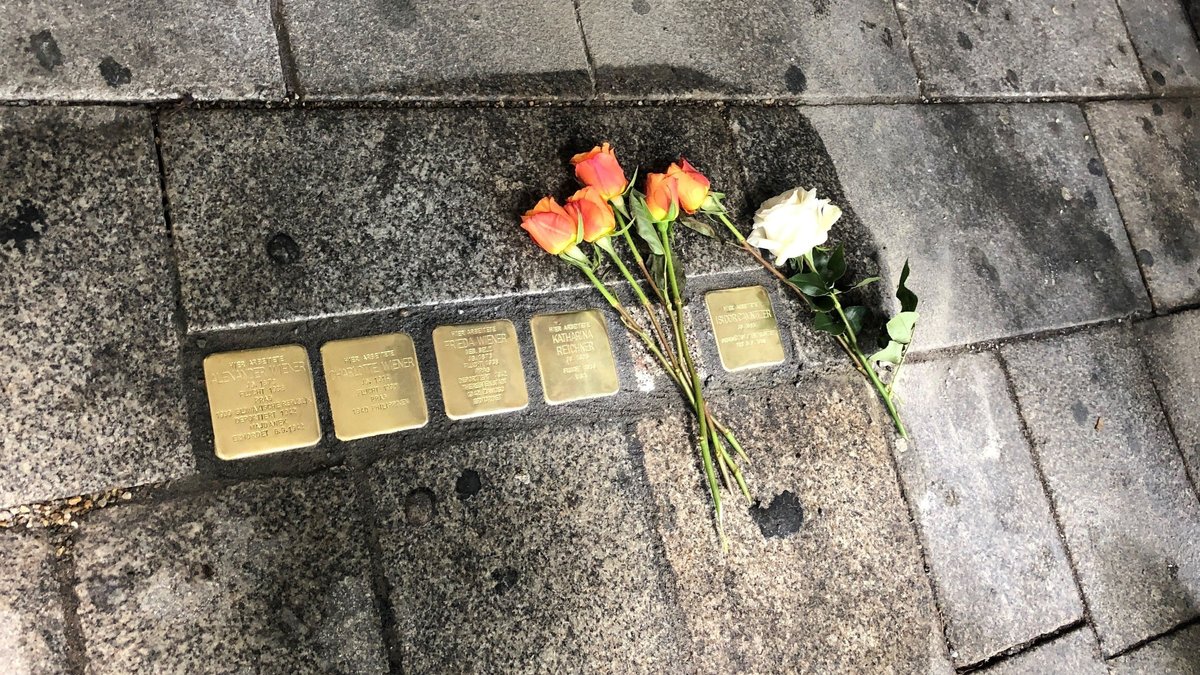 Stolpersteine, in den Boden eingelassene, vergoldete Steine mit Daten verfolgter und ermordeter Opfer des Nationalsozialismus. Neben den Steinen liegen mehrere rot-gelbe Rosen und eine weiße Rose.