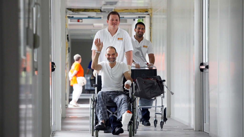 Patient mit Arm und Bein in Gips wird in einem Rollstuhl von einer Pflegekraft durch einen langen Gang entlang des Klinikgebäudes geschoben.