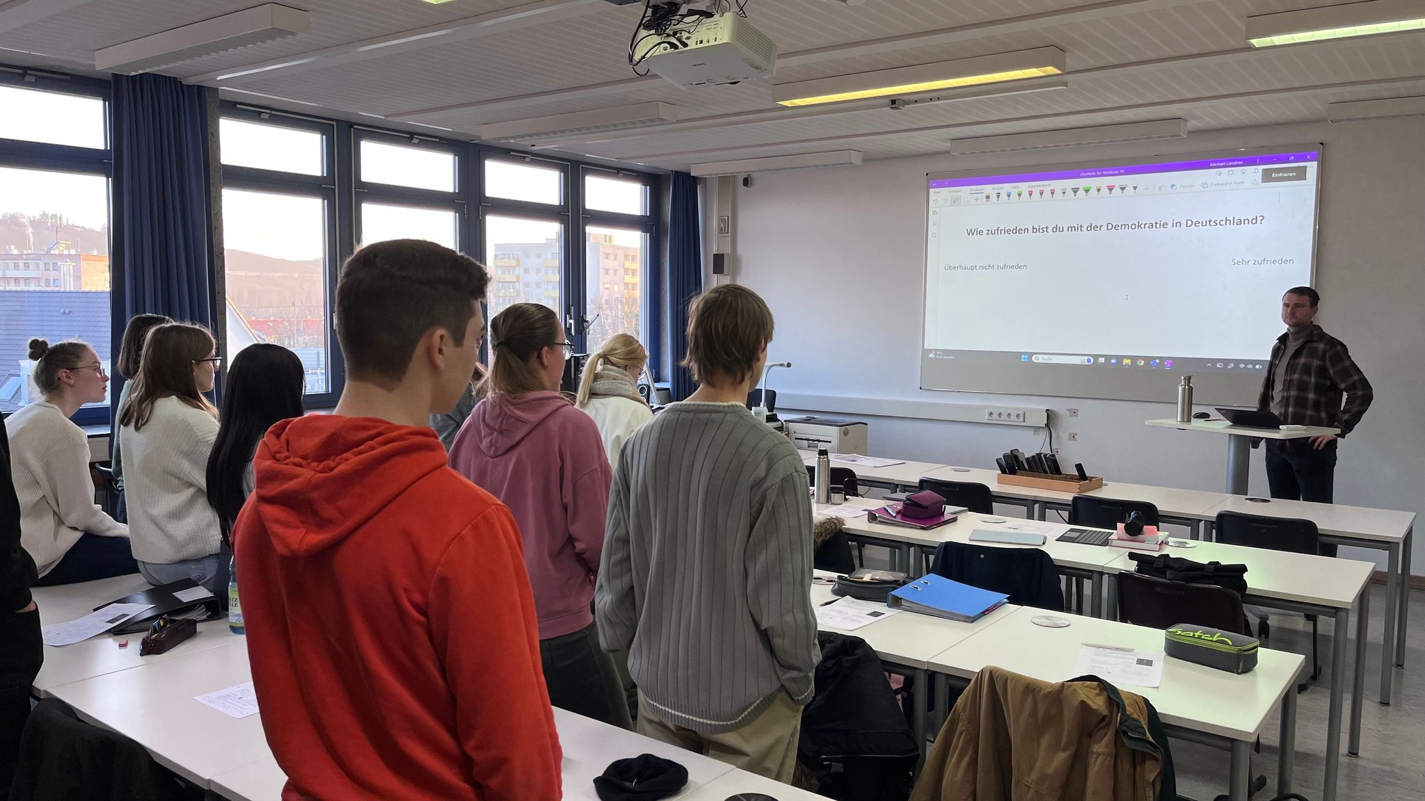 Schüler stehen im Klassenzimmer verteilt und schauen an die digitale Tafel.