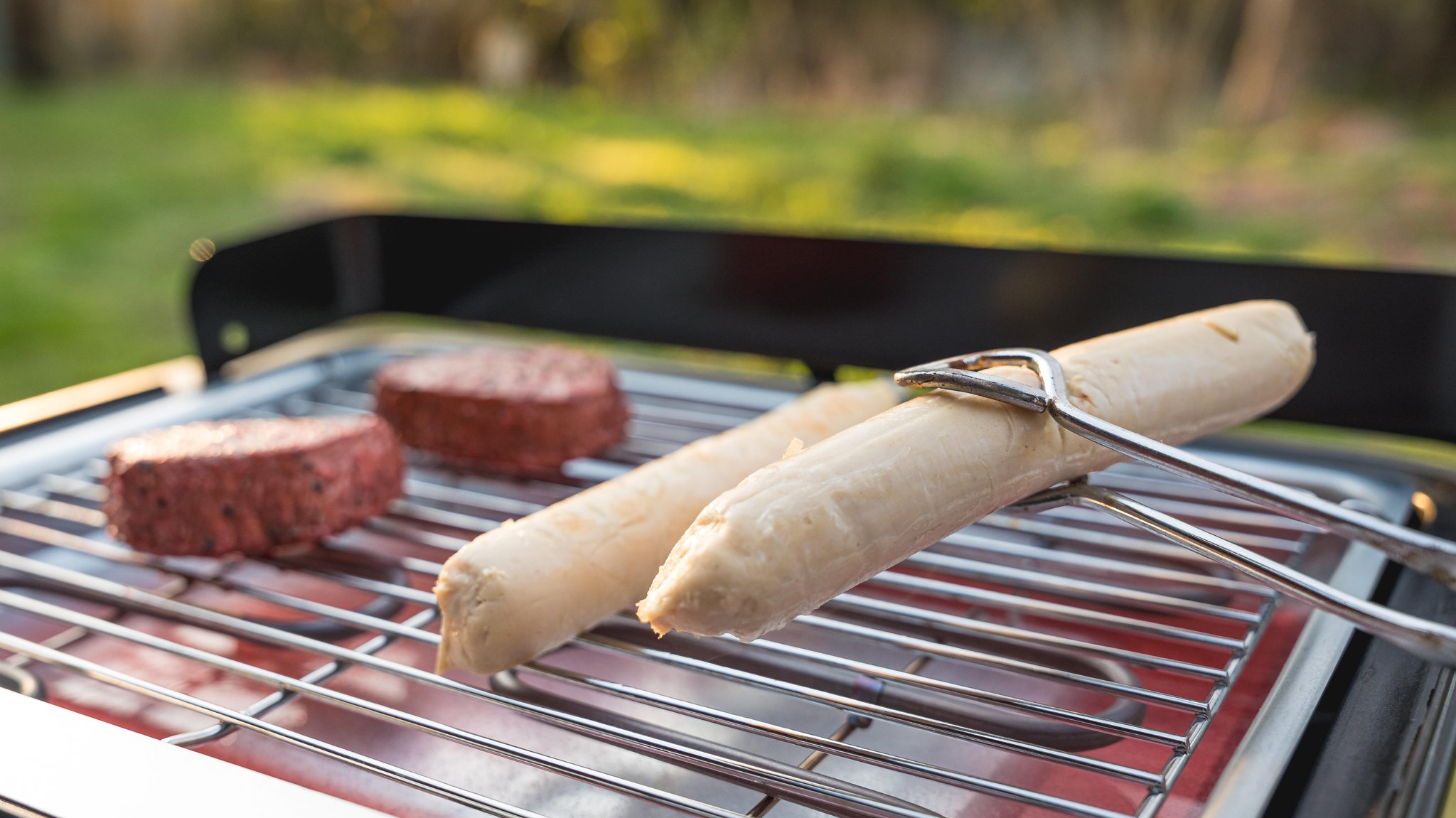 Eine vegane Wurst wird von einer Grillzange hochgehalten und fleischfreie Frikadellen liegen auf einem Grill. | Bild: picture alliance / CHROMORANGE | Michael Bihlmayer Eine vegane Wurst wird von einer Grillzange hochgehalten und fleischfreie Frikadellen liegen auf einem Grill.