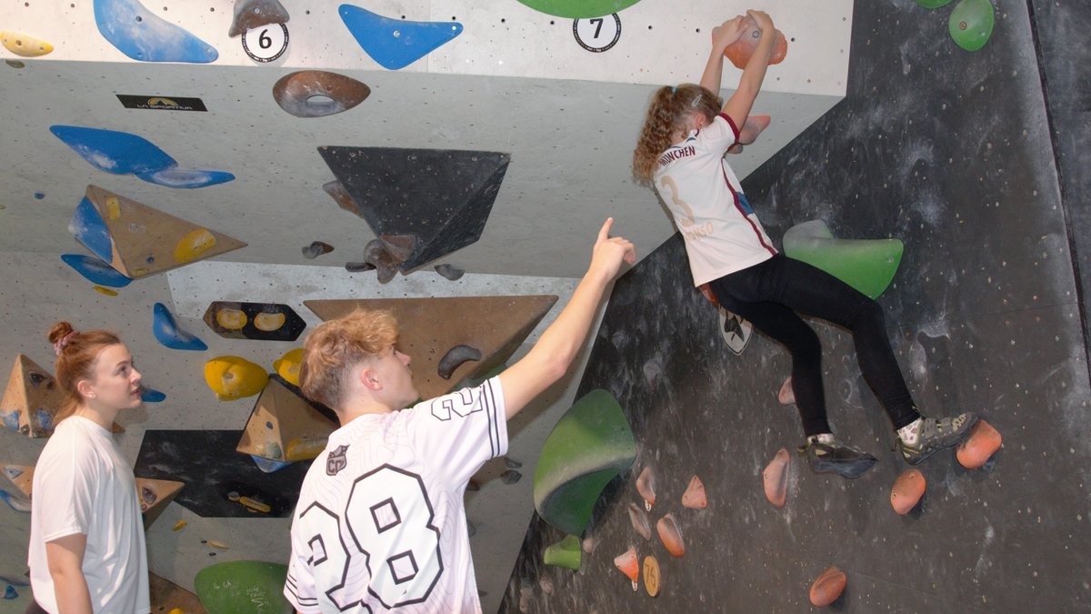 Ein Mädchen bouldert an der Wand nach oben, während ihr zwei Jugendliche zuschauen.