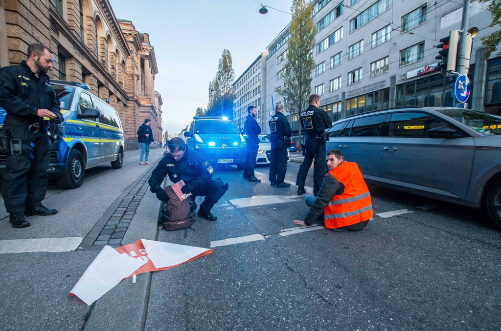 07.11.22: Ein mit der Hand auf der Straße festgeklebter Klima-Aktivist, umgeben von Polizisten.