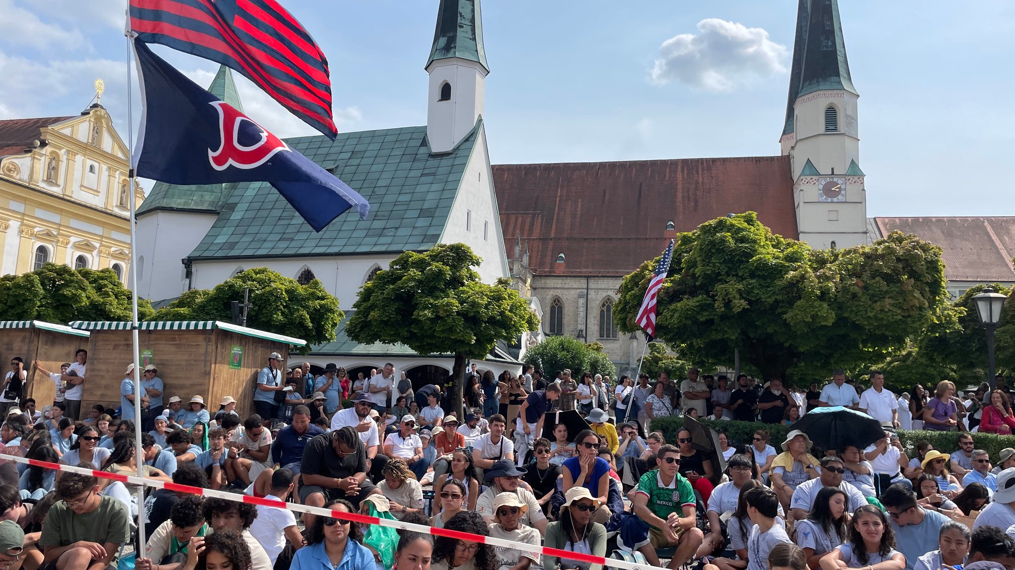Der Kapellplatz in Altötting mit Kirche und Gnadenkapelle, davor eine Menschenmenge im Freien und zwei Fahnen aus den USA. Auf dem Kapellplatz in Altötting feierten 7.000 Gläubige aus den USA den Abschluss ihrer Pilgerreise.