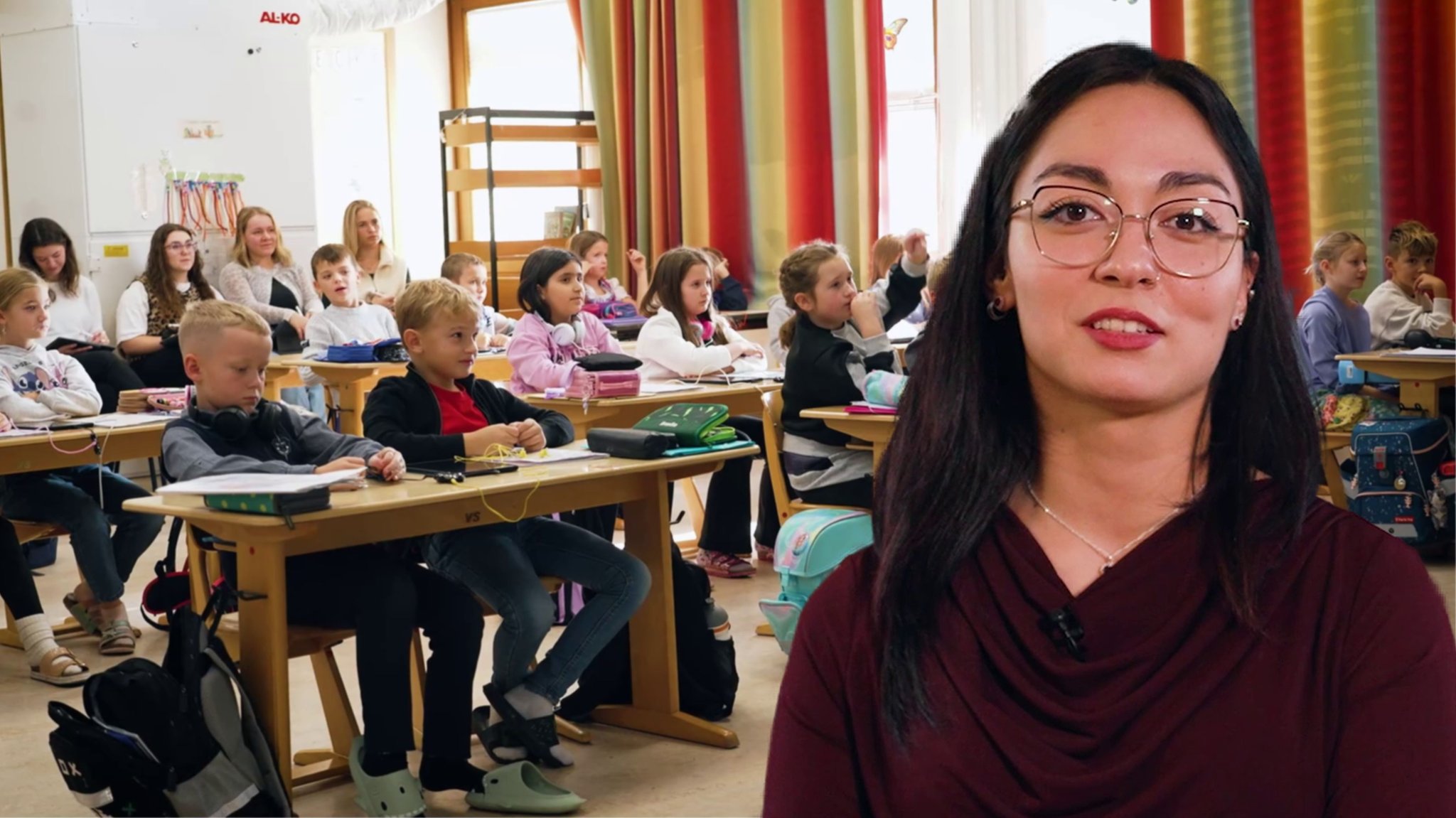 Ebru im Portrait vor einem Klassenraum mit Kindern. Sie ist Lehramtsanwärterin im Vorbereitungsdienst an der Grundschule Burgkirchen an der Alz. Sie unterrichtet Englisch im Hauptfach und Deutsch, Mathematik und Kunst im Nebenfach. 