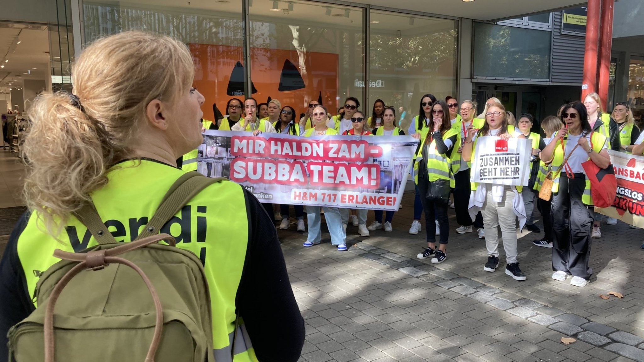 Streikende in gelben Streikwesten mit Transparenten vor einem Geschäft.