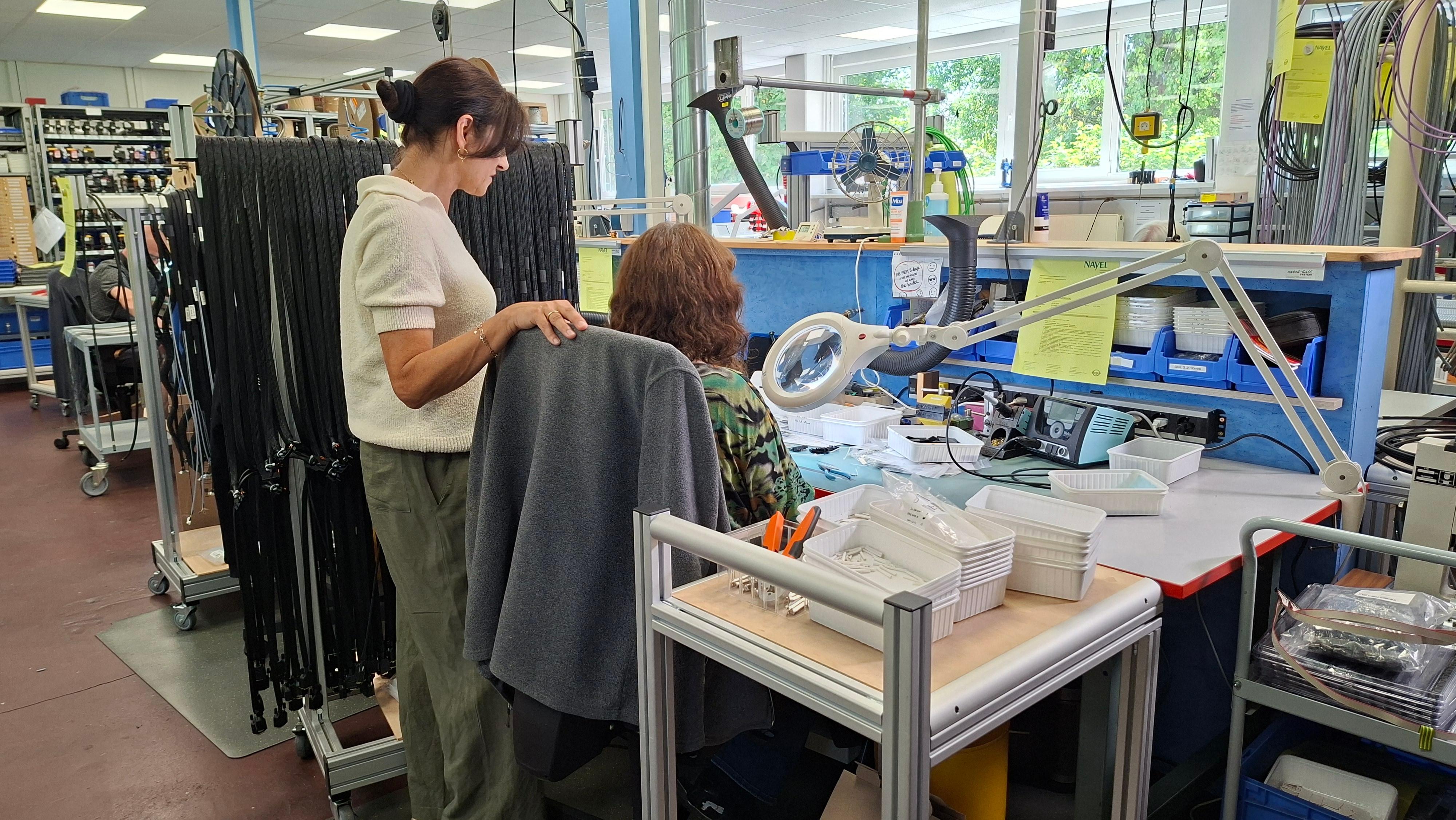 An einem Arbeitsplatz konfektioniert eine Frau Kabel, dahinter eine weitere Frau, die ihr zuschaut.
