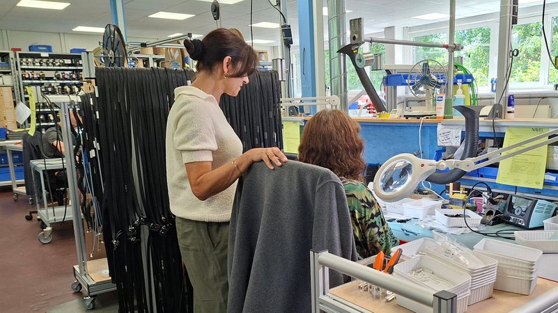 An einem Arbeitsplatz konfektioniert eine Frau Kabel, dahinter eine weitere Frau, die ihr zuschaut. | Bild: BR/Eleonore Birkenstock An einem Arbeitsplatz konfektioniert eine Frau Kabel, dahinter eine weitere Frau, die ihr zuschaut.