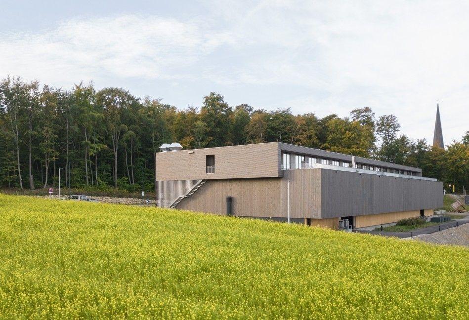 Im Vordergrund eine blühende Wiese, im Mittelgrund ein Flachbau mit Wohnungs-Etage und im Hintergrund ein Kirchturm