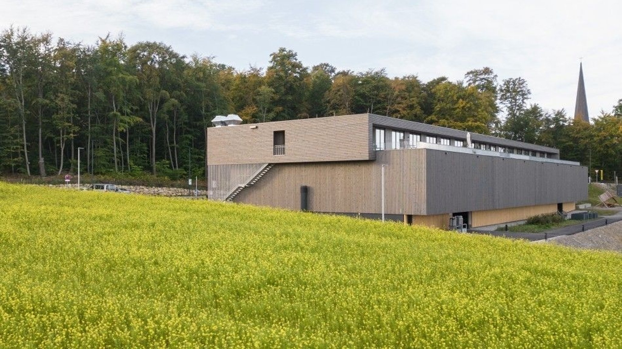 Im Vordergrund eine blühende Wiese, im Mittelgrund ein Flachbau mit Wohnungs-Etage und im Hintergrund ein Kirchturm