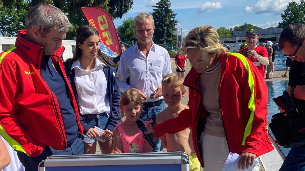 Markus Söder steht mit Kindern am Freibad in Nürnberg. Er trägt eine rote DLRG-Jacke.